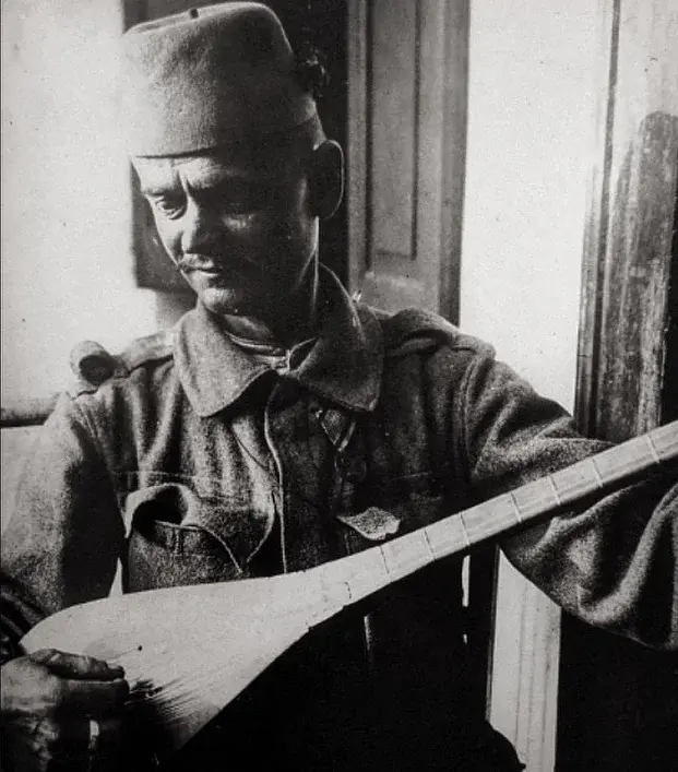 Un vieux fantassin bosno-herzégovinien joue de la šargija en rythme et musique, un luth traditionnel à long manche, sur un champ de bataille européen pendant la Première Guerre mondiale.