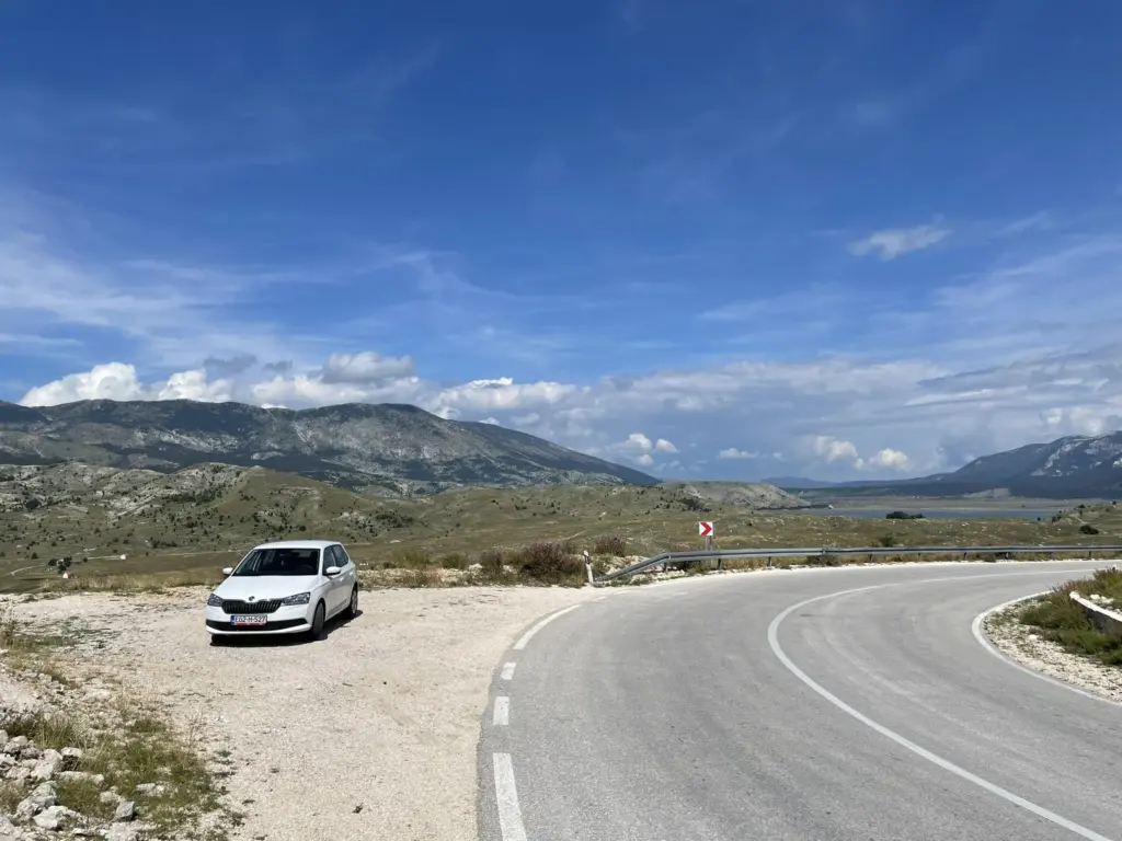 Voiture garée sur le bas-côté d’une route sinueuse en Bosnie, avec un paysage de collines et de forêts en arrière-plan.