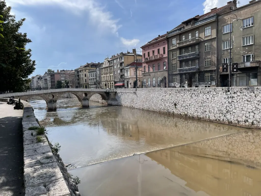 Vue depuis le Pont Latin à Sarajevo, avec la rivière Miljacka, les façades anciennes et les collines environnantes sous un ciel clair.