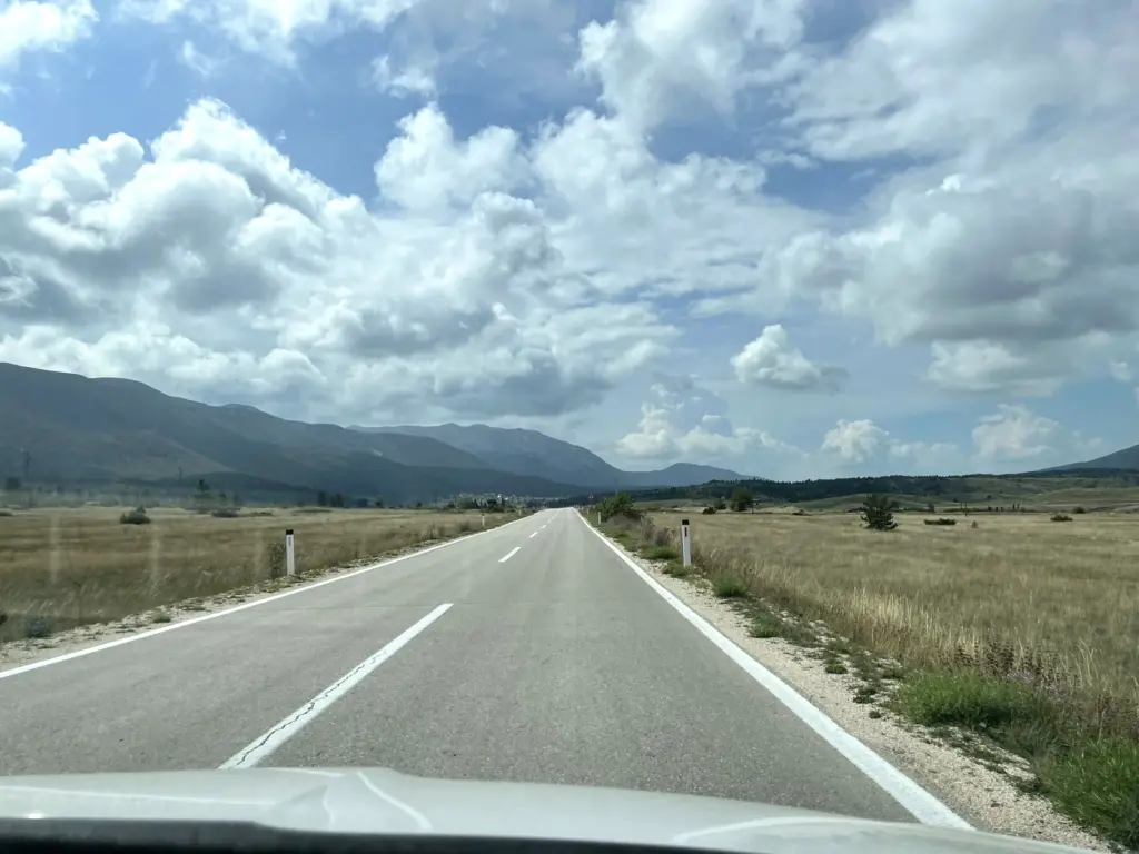 Route sinueuse traversant le parc naturel de Blidinje, encadrée par des montagnes majestueuses au loin sous un ciel clair.